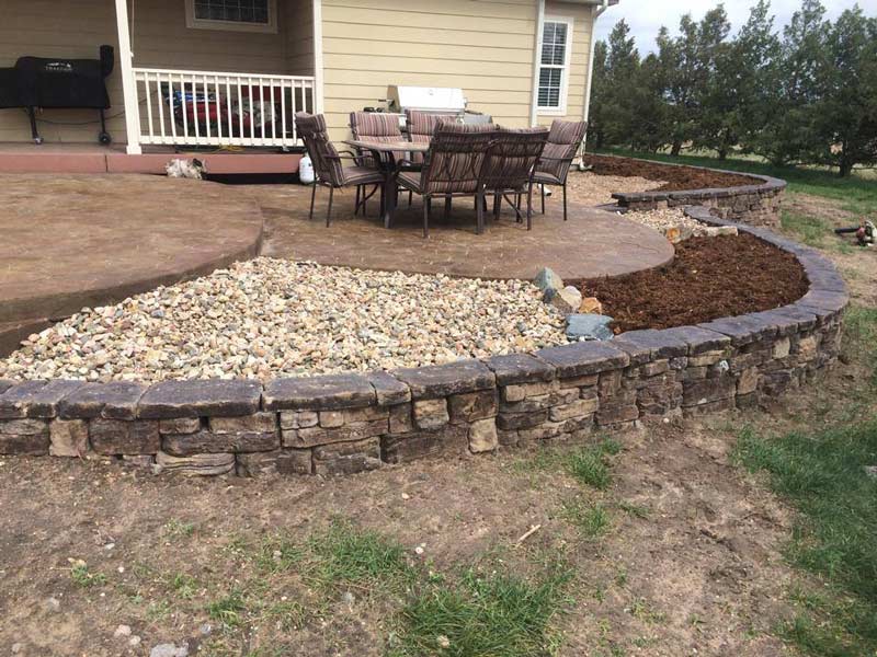 An open stone patio with three sections of open space filled with pebbles or mulch in front of a backyard created by by Total Lawn Care & Landscape in Sterling, CO