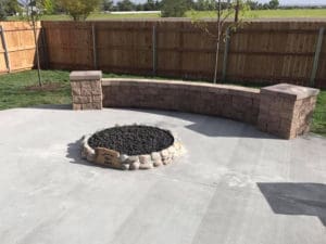 A small built in fire pit in front of a small stone wall in front of a wooden fence and a couple trees planted by Total Lawn Care & Landscape in Sterling, CO