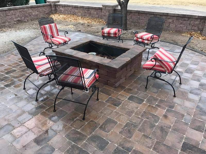 A few metal chairs with cushions all arranged around a built in fire place in a stone patio deisgned by Total Lawn Care & Landscape in Sterling, CO