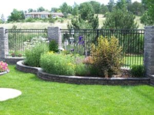 A flower bed bursting with large bushes and flowers in front of a black metal fence within a small stone wall by Total Lawn Care & Landscape in Sterling, CO