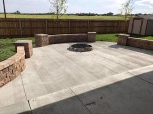 A small built in fire pit in the middle of a paved patio with three small stone walls alongside a lawn designed and created by Total Lawn Care & Landscape in Sterling, CO