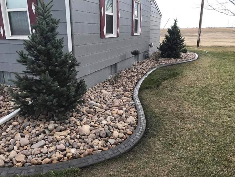 A stone siding flower bed with two medium sized trees next to a patchy grassy lawn landscaped by Total Lawn Care & Landscape in Sterling, CO