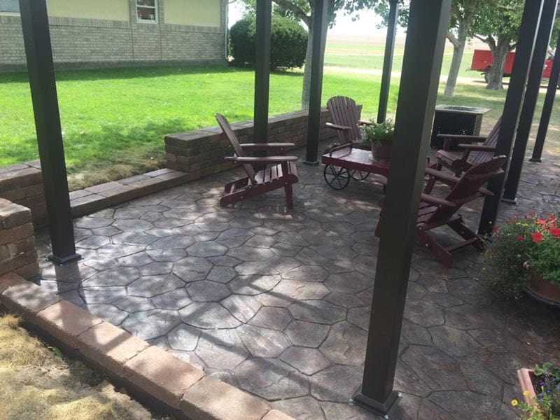 Four wooden lounge chairs on a covered stone patio with metal poles and some flowers and other decorative elements installed by Total Lawn Care & Landscape in Sterling, CO