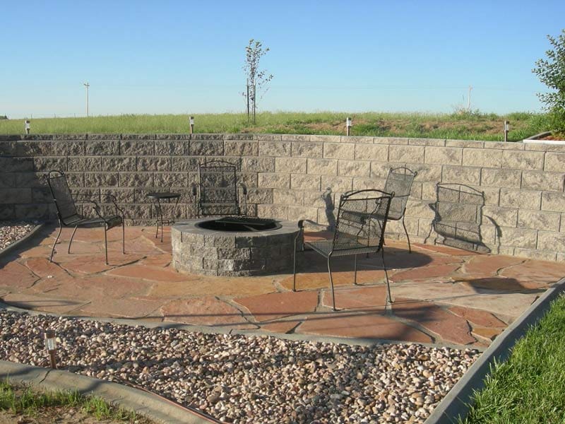 Four metal chairs surrounding a built in fire pit by Total Lawn Care & Landscape in Sterling, CO with a large stone wall keeping a wall of dirt and grass separated